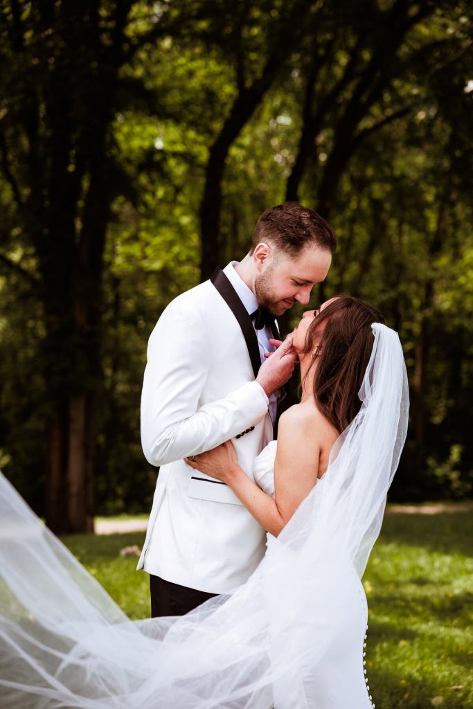 A bride and groom share a romantic moment outdoors, surrounded by greenery. The bride wears a strapless white gown and a long veil, while the groom is dressed in a formal white suit with a black bow tie.