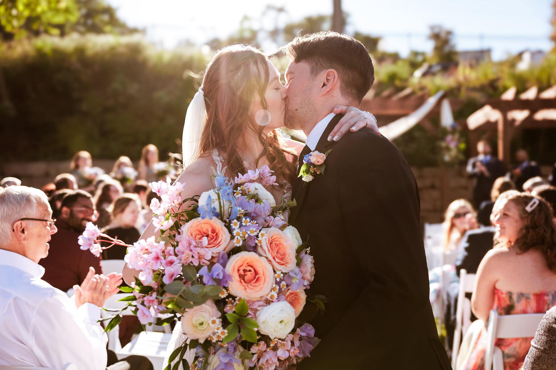 bride and groom kiss in the aisle holding flowers while the sun flares at the camera lens